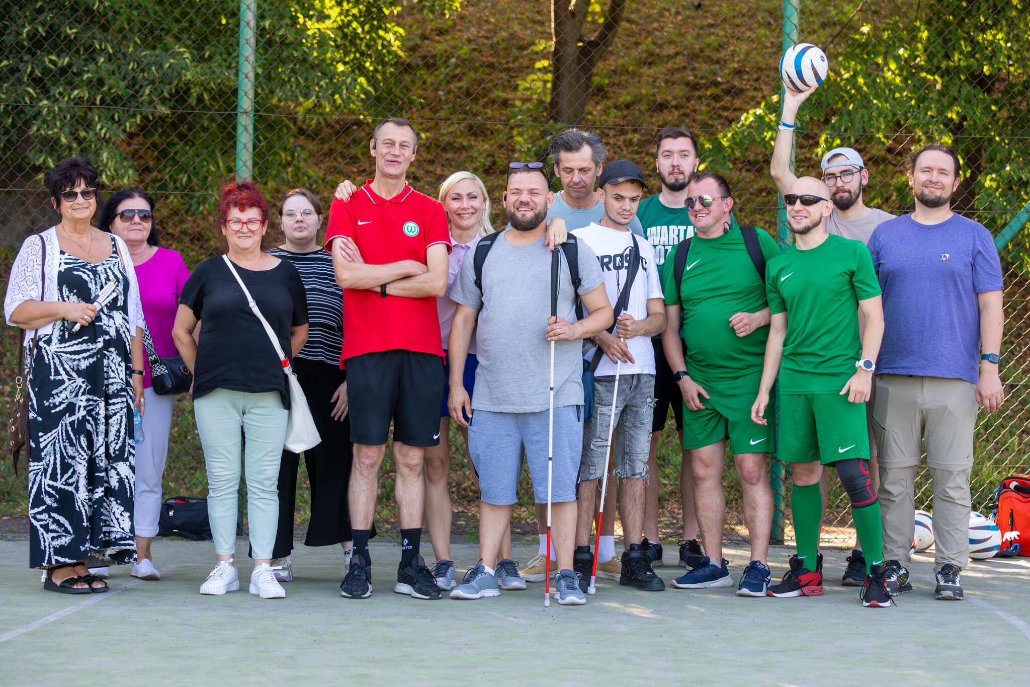 Grupa uczestników i organizatorów pozuje do wspólnego zdjęcia na boisku sportowym. Wśród nich są osoby z białymi laskami oraz zawodnicy ubrani w zielone stroje sportowe. W tle znajduje się ogrodzenie i drzewa, a jedna z osób trzyma nad głową piłkę.