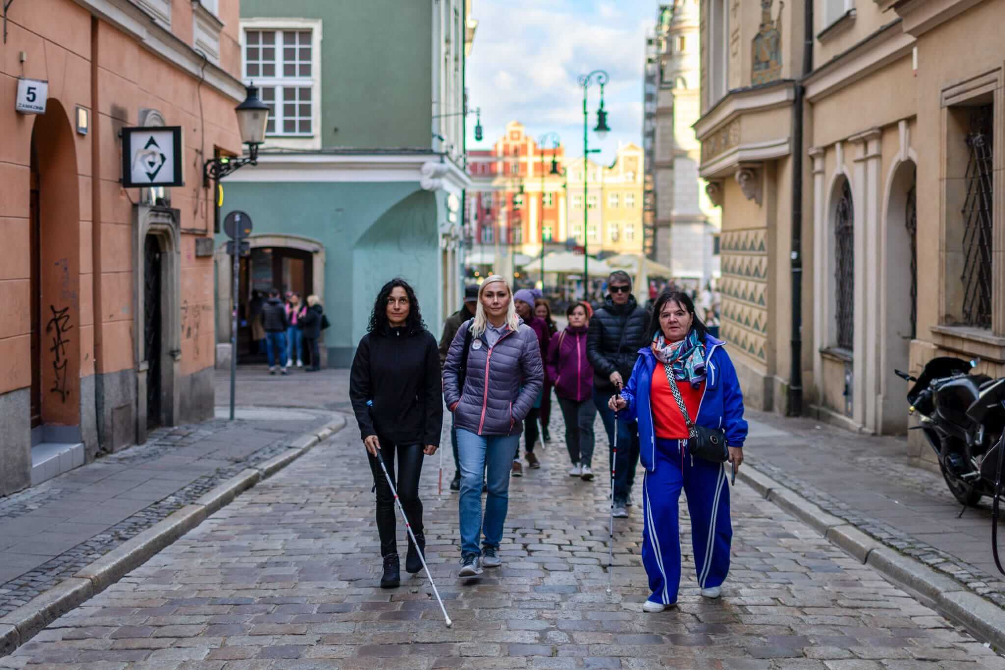 Grupa osób, w tym uczestnicy z białymi laskami, stoi na brukowanej ulicy poznańskiej Starówki. W tle widoczne są kolorowe kamienice oraz zabytkowa zabudowa. Fotografia dokumentuje wspólne zwiedzanie miasta przez osoby z niepełnosprawnością wzroku i ich towarzyszy.