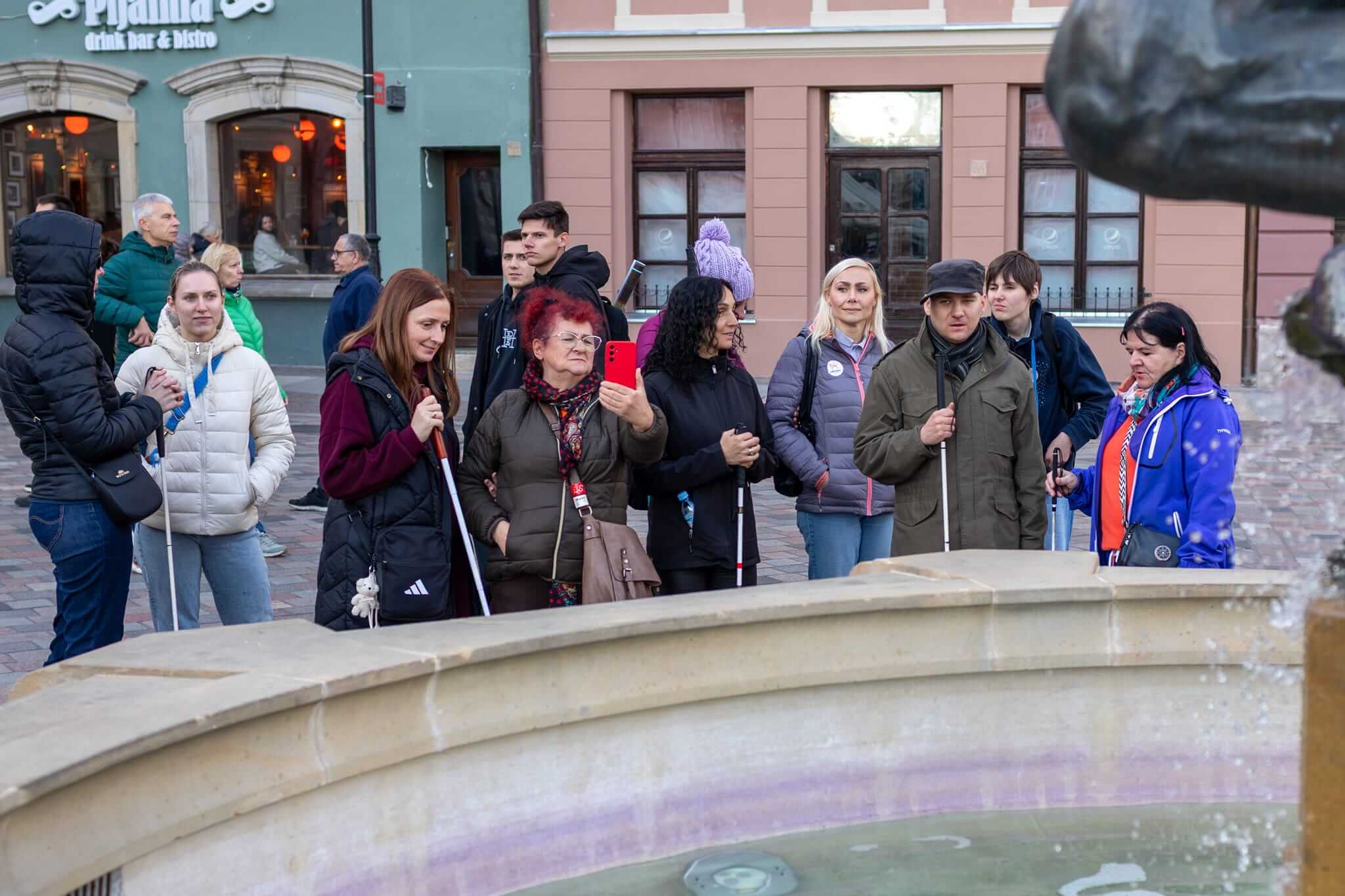 Grupa uczestników spaceru stoi przy kamiennej fontannie na poznańskim Starym Rynku. Jedna z kobiet trzyma telefon, a kilka osób korzysta z białych lasek. Fotografia przedstawia chwilę odpoczynku i rozmowy podczas wspólnego zwiedzania miasta.