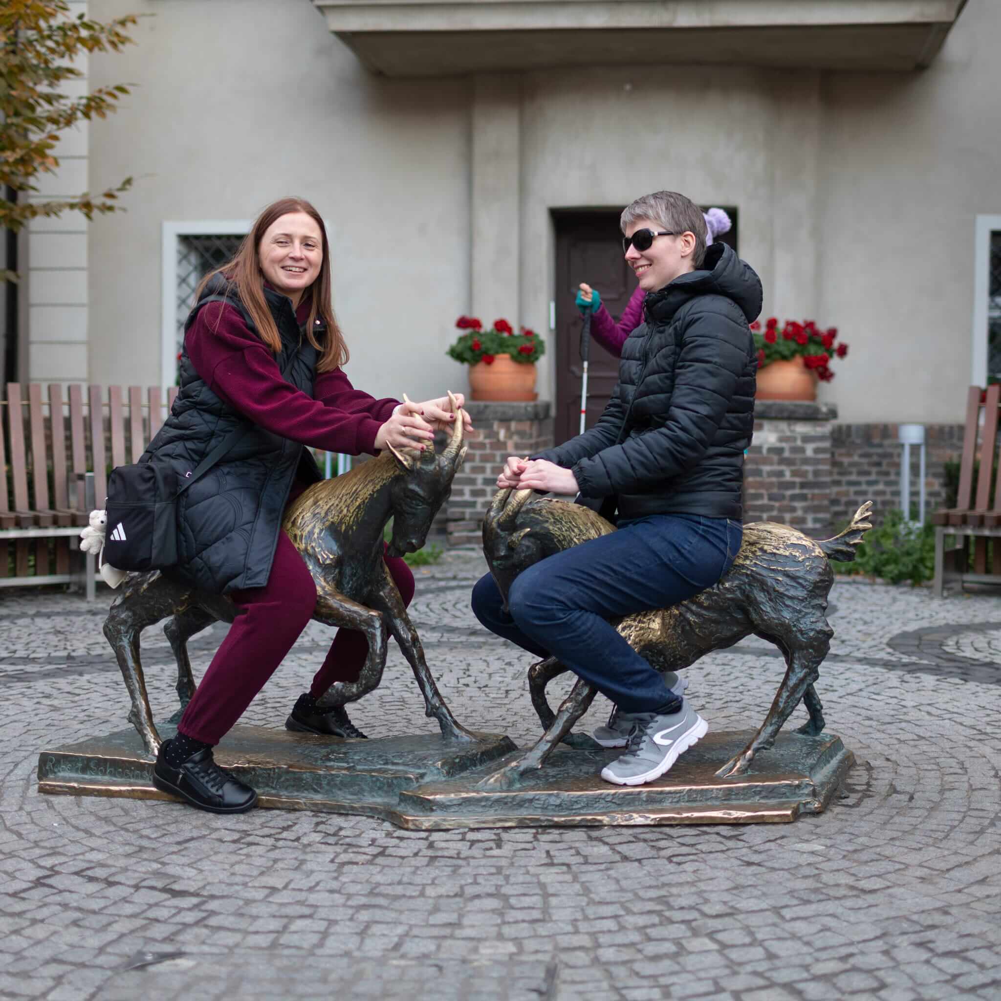 Dwie kobiety pozują przy metalowych rzeźbach koziołków na poznańskiej Starówce. Jedna z nich dotyka rogów rzeźby, a druga siedzi na grzbiecie drugiego koziołka. Zdjęcie przedstawia uczestniczki spaceru integracyjnego po Poznaniu.