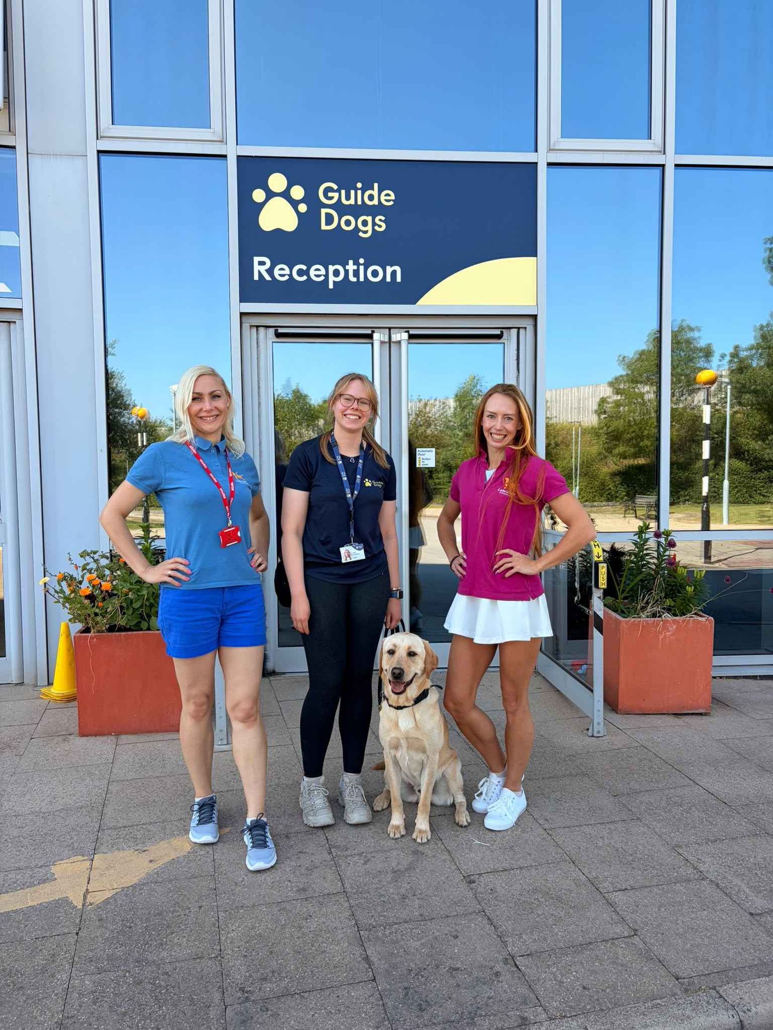 Trzy uśmiechnięte kobiety stoją przed wejściem do ośrodka szkoleniowego Guide Dogs pod tablicą z napisem „Guide Dogs Reception”. Pomiędzy nimi siedzi spokojnie żółty labrador. Wszystkie patrzą w stronę aparatu, a w tle widać przeszkloną fasadę budynku oraz donice z kwiatami. Zdjęcie dokumentuje wizytę w brytyjskim ośrodku szkolenia psów przewodników i wspólne spotkanie z jego pracownikami.