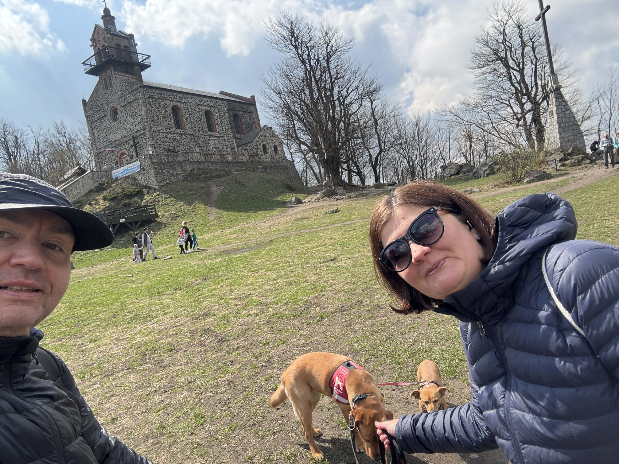 Para opiekunów stoi na tle kamiennego kościoła położonego na wzgórzu, obok dwóch brązowych psów w&nbsp;trakcie szkolenia. Jednego z&nbsp;psów, ubranego w&nbsp;czerwoną kamizelkę Fundacji Labrador, widać podczas eksplorowania terenu na smyczy. Zdjęcie przedstawia wspólną wycieczkę socjalizacyjną, podczas której psy poznają nowe miejsca, zapachy i&nbsp;otoczenie, przygotowując się do pracy w&nbsp;zróżnicowanych warunkach.