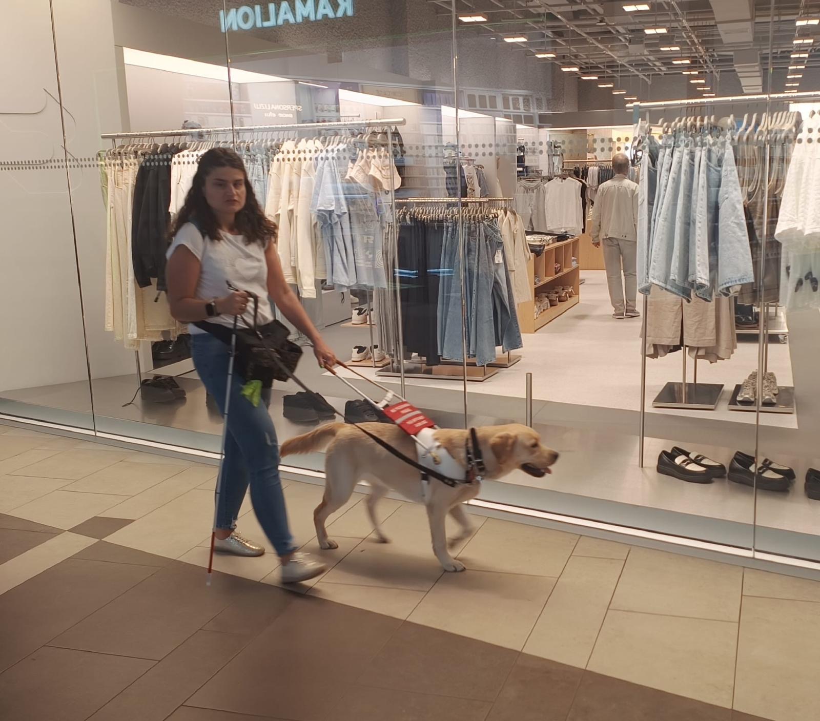 Kobieta z&nbsp;białą laską idzie przez galerię handlową z&nbsp;jasnym labradorem przewodnikiem w&nbsp;uprzęży. Mijają przeszklony sklep z&nbsp;ubraniami, za którego witryną stoją wieszaki z&nbsp;odzieżą.