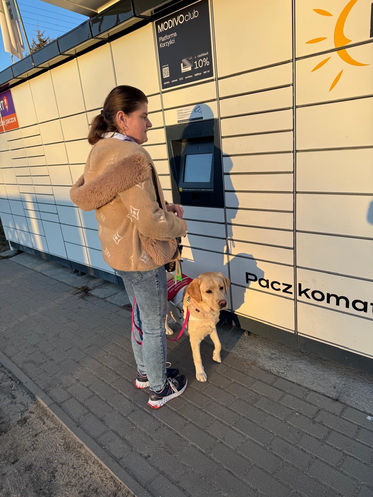Kobieta stoi przed paczkomatem, a&nbsp;obok niej czeka jasny labrador przewodnik w&nbsp;uprzęży. Pies stoi spokojnie przy nodze opiekunki, skierowany w&nbsp;stronę aparatu.