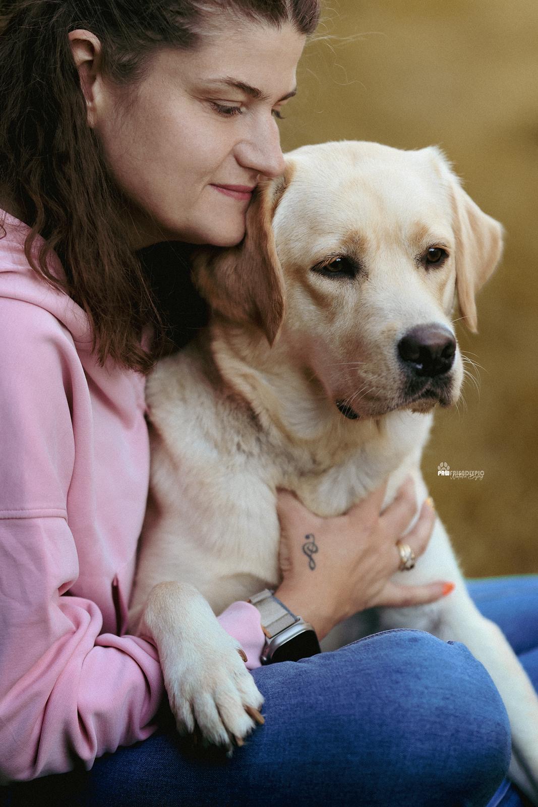 Zbliżenie przedstawia kobietę przytulającą jasnego labradora. Kobieta opiera twarz o&nbsp;głowę psa i&nbsp;obejmuje go obiema rękami.