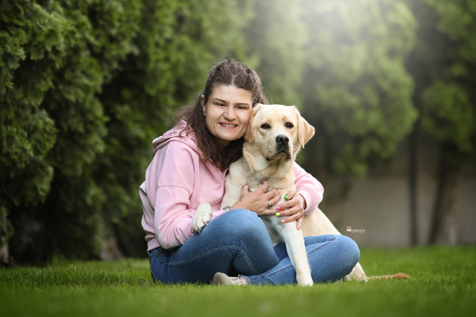 Kobieta siedzi na trawie i&nbsp;obejmuje jasnego labradora. Oboje patrzą w&nbsp;stronę aparatu, a&nbsp;w tle widać rozmyte drzewa.