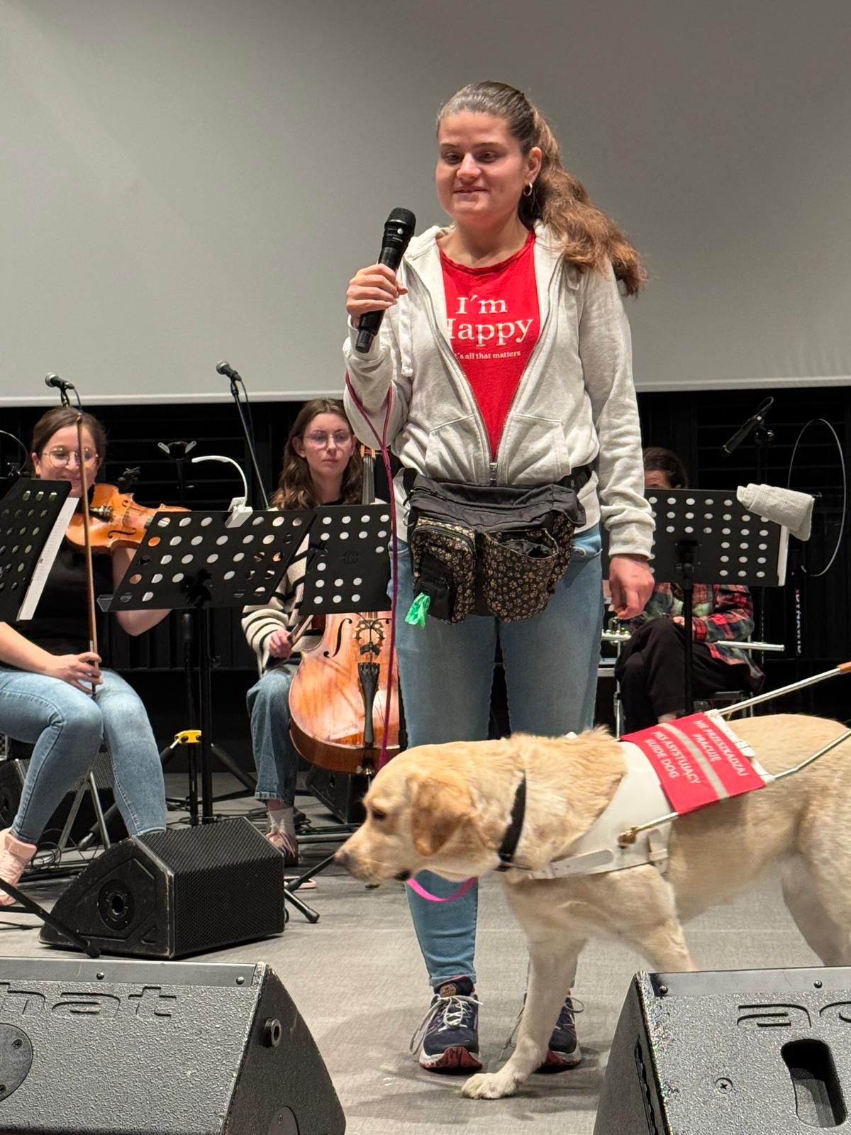 Kobieta stoi na scenie z&nbsp;mikrofonem w&nbsp;dłoni. Obok niej znajduje się jasny labrador przewodnik w&nbsp;uprzęży, a&nbsp;w tle siedzą muzycy z&nbsp;instrumentami i&nbsp;pulpitami do nut.
