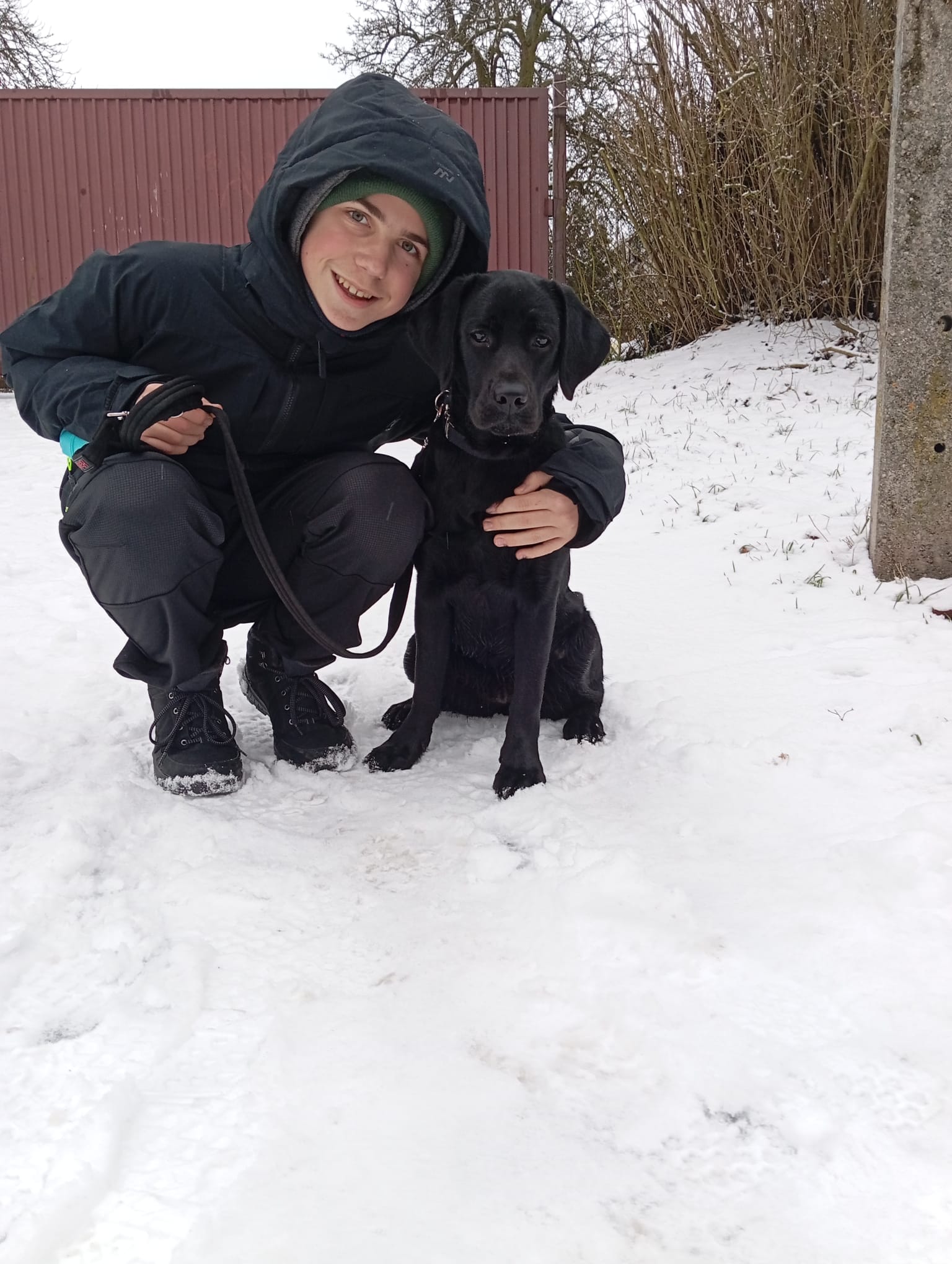 Dziecko w&nbsp;zimowym ubraniu kuca na śniegu obok czarnego labradora siedzącego spokojnie na smyczy. Jedną ręką obejmuje psa, a&nbsp;drugą trzyma smycz. W&nbsp;tle widać ośnieżone podwórko, ogrodzenie i&nbsp;bezlistne krzewy. Oboje patrzą w&nbsp;stronę aparatu.
