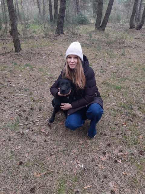 Osoba w&nbsp;czarnej kurtce puchowej i&nbsp;białej czapce kuca w&nbsp;lesie, obejmując czarnego labradora retrievera. Wokół widać sosnowy las z&nbsp;igliwiem i&nbsp;szyszkami na ziemi. Pochmurna, jesienna lub wczesnowiosenna pogoda