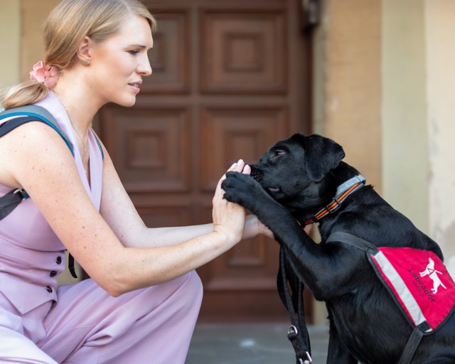 Czarny labrador retriever w&nbsp;czerwonej kamizelce Fundacji Labrador podaje łapę osobie kucającej naprzeciwko niego. Osoba ma na sobie jasnofioletową sukienkę i&nbsp;plecak. Za nimi widać ciemne, drewniane drzwi. Zdjęcie zrobione w&nbsp;ciepły dzień.